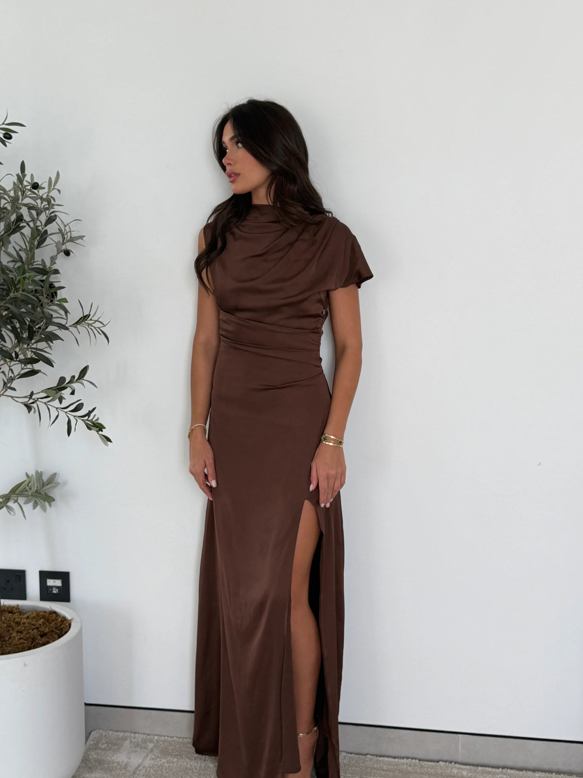 Brown satin maxi dress with a draped neckline, worn against a white wall